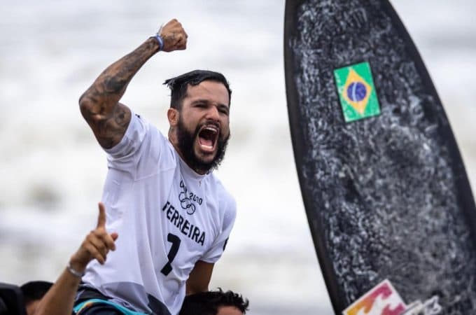 Na imagem, o surfista Ítalo Ferreira, de branco, ergue o punho direito e grita. Ele entrou para a história dos medalhistas brasileiros em jogos Olímpicos e é erguido por pessoas de sua equipe técnica na aria de uma praia no Japão.