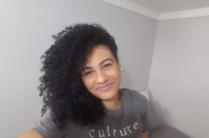 Raquel, no centro da imagem, sorri. Ela é uma mulher negra, tem os cabelos cacheados e usa camisa cinza.