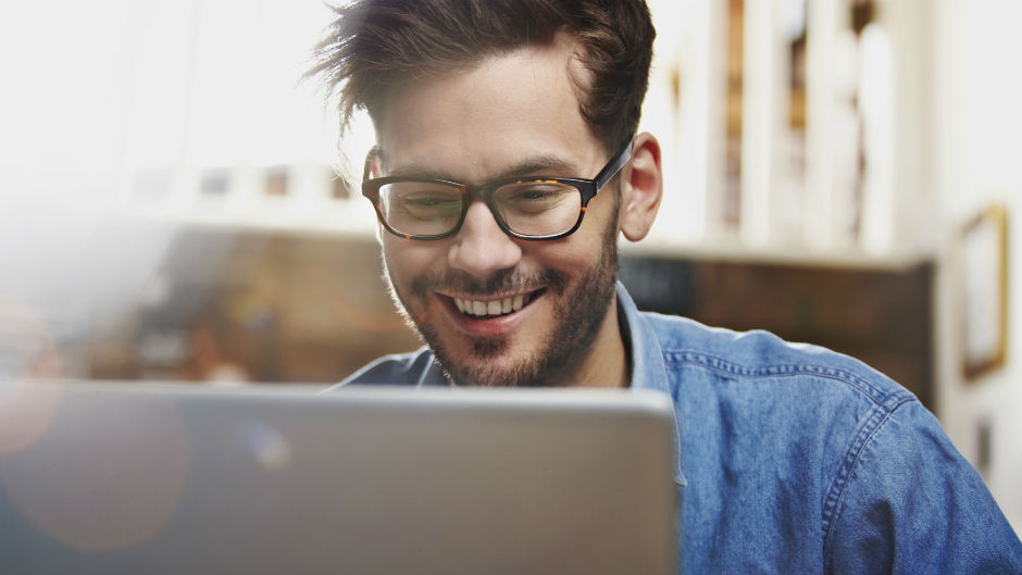 Homem jovem de óculos sorri em frente ao laptop