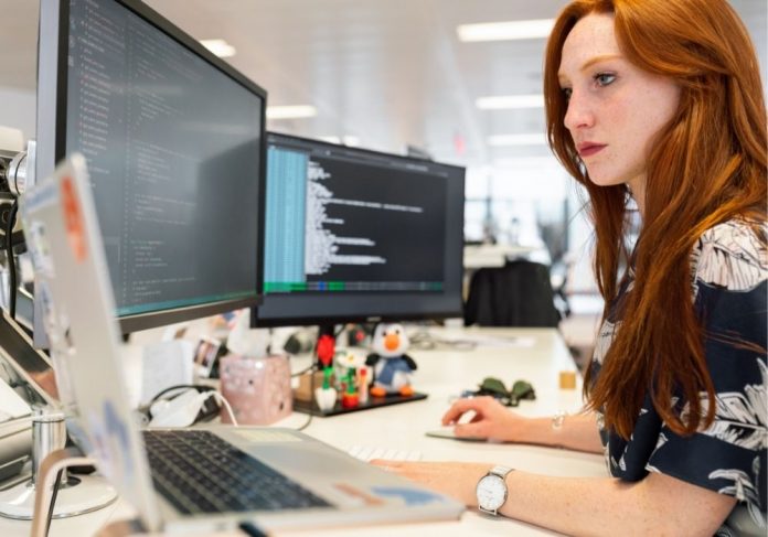 mulher ruiva olhando para duas telas de computador em que estão gráficos e números. ela representa uma profissional de data science