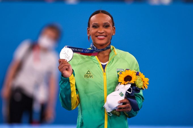 Rebeca Andrade é uma mulher jovem, negra, que usa o uniforme do Brasil durante a cerimônia de premiação das Olímpiadas de Tóquio. Ela exibe uma medalha de prata e carrega um buquê de flores no centro da imagem, em destaque.