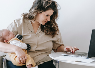 Licença-maternidade: mulher digita em um notebook com a mão esquerda, enquanto segura um bebe com a mão direita