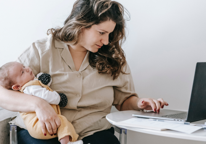Licença-maternidade: mulher digita em um notebook com a mão esquerda, enquanto segura um bebe com a mão direita