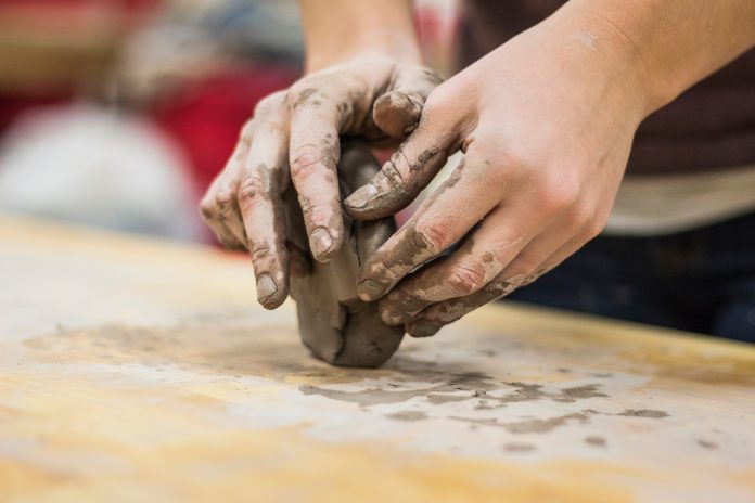 Um par de mãos brancas modela uma massa de argila em sobre uma mesa de madeira. A imagem representa uma pessoa com perfil hands on