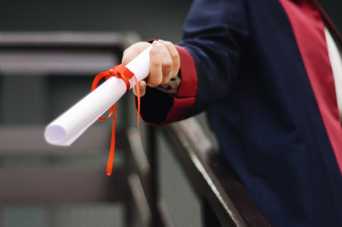 Uma pessoa com uma beca de formatura estende seu diploma, que está em destaque na imagem. Acesse o Guia de Profissões