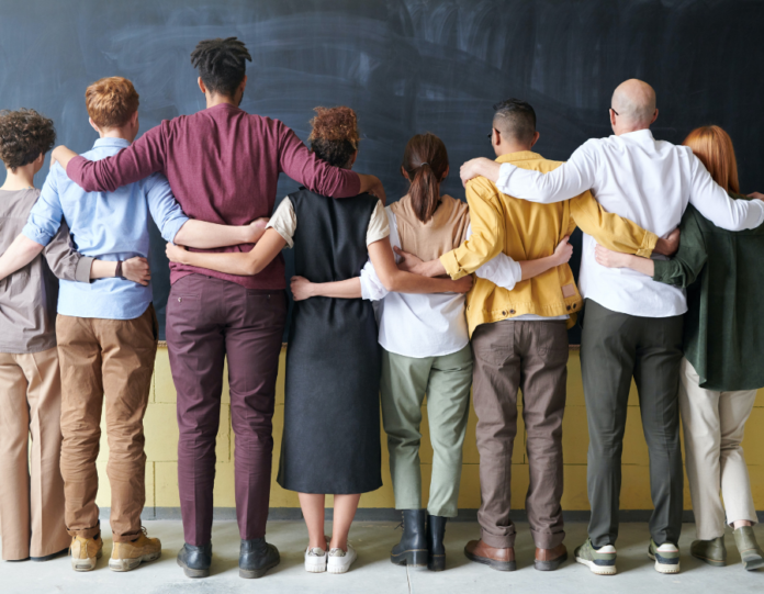 pessoas de diferentes etnias abraçadas, de costas, em uma sala de aula representando o ideal de diversidade de inclusão